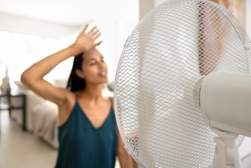 Žena se dotýká čela, čelí elektrickému ventilátoru, cítí úlevu od letního tepla, ochlazuje se během teplého dne, bojuje s vysokou teplotou v interiéru bez klimatizace, klimatizace